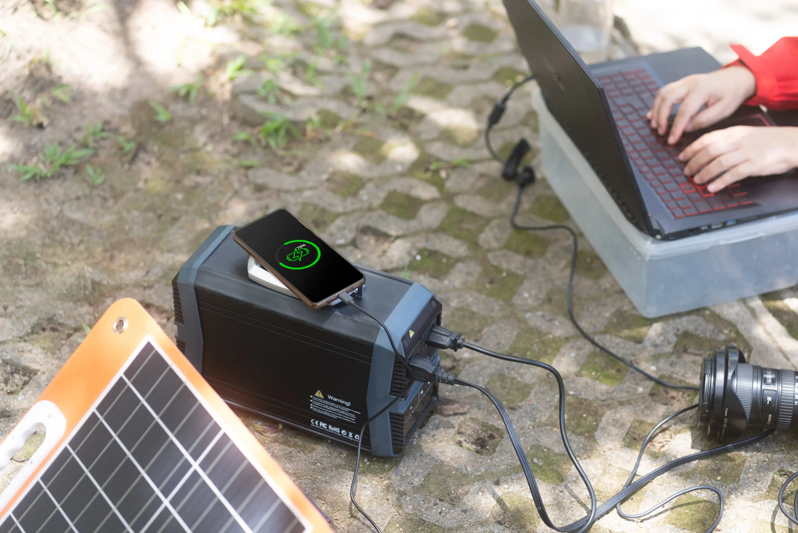 fsebattery portable power station simultaneously charging smartphone and laptop on stone pavement in outdoor setting. Solar panel positioned nearby demonstrating sustainable energy solution for mobile productivity with green charging indicators visible on phone screen.