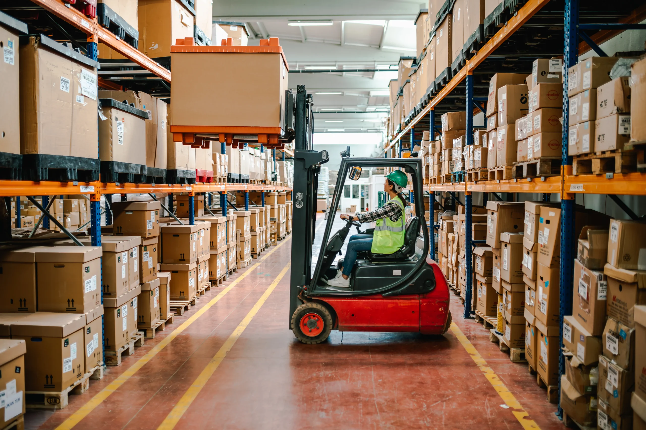 Safety-vested worker driving red forklift with large orange crate in industrial warehouse, powered by fsebattery lithium batteries. Background of organized orange/blue shelves with palletized goods under bright facility lighting.