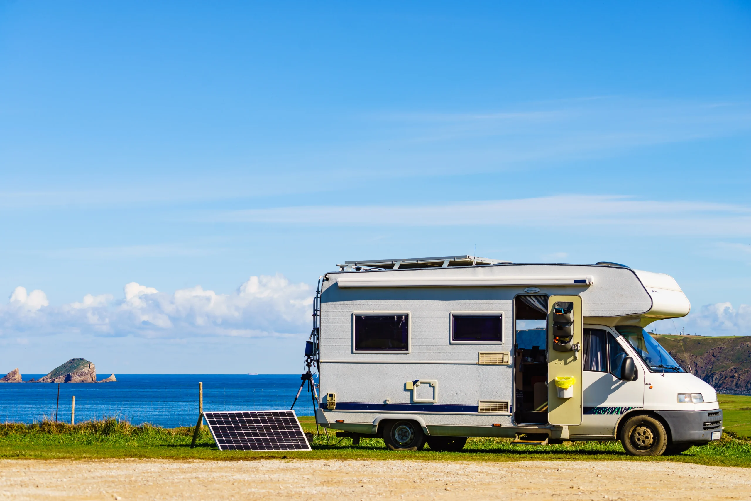 White RV with blue accents parked on coastal grassland, door open revealing interior space powered by fsebattery LiFePO4 battery systems. Roof-mounted solar panels charging storage units for off-grid living with turquoise sea and islands in background.