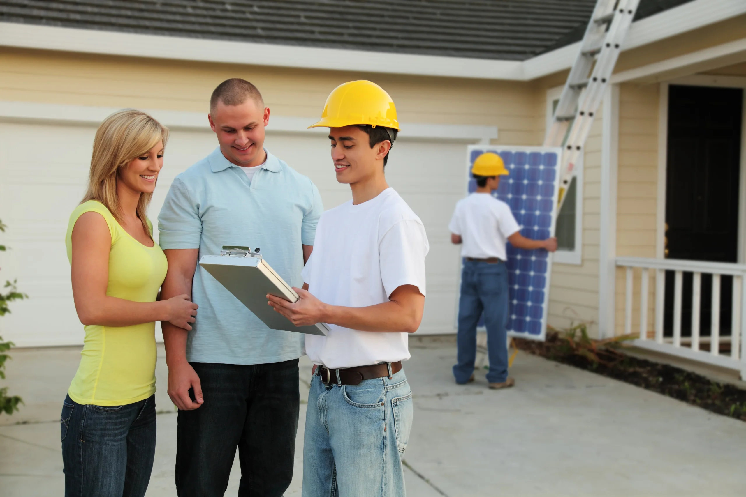 Homeowner couple consulting with safety-helmeted technician holding clipboard, while background worker installs solar panels on beige house with garage door and ladder. Demonstrates professional residential solar installation process.